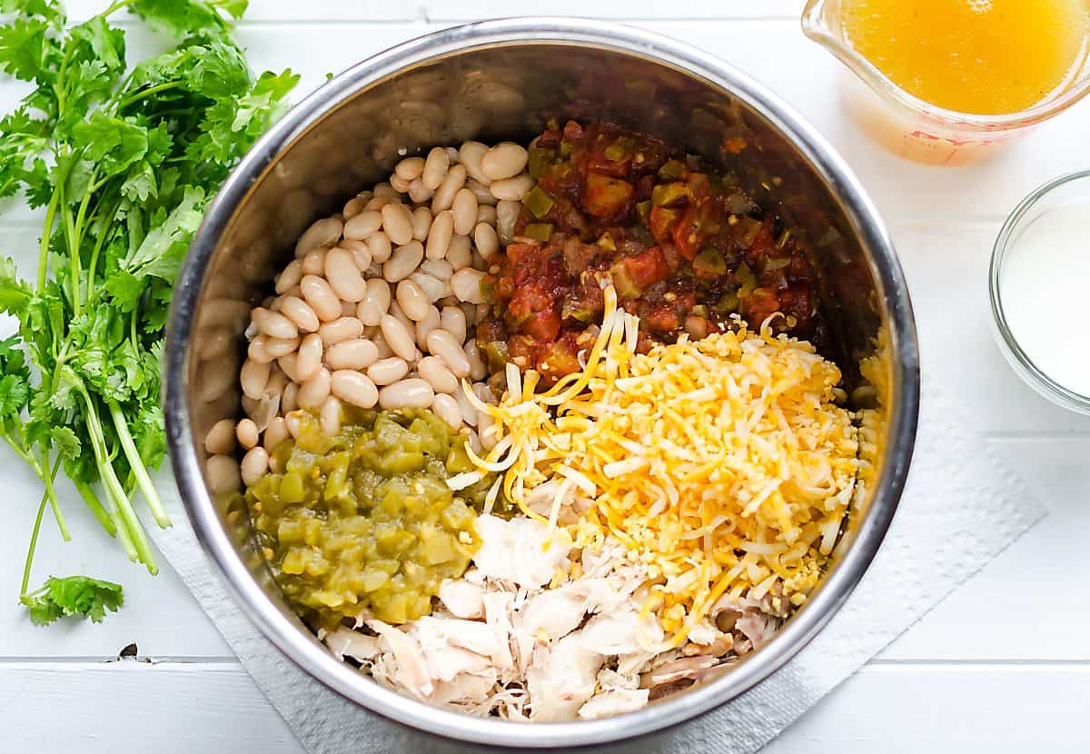 crockpot/instant pot filled with white chicken chili ingredients and segmented-white beans, salsa, green chilies, chicken and cheeese