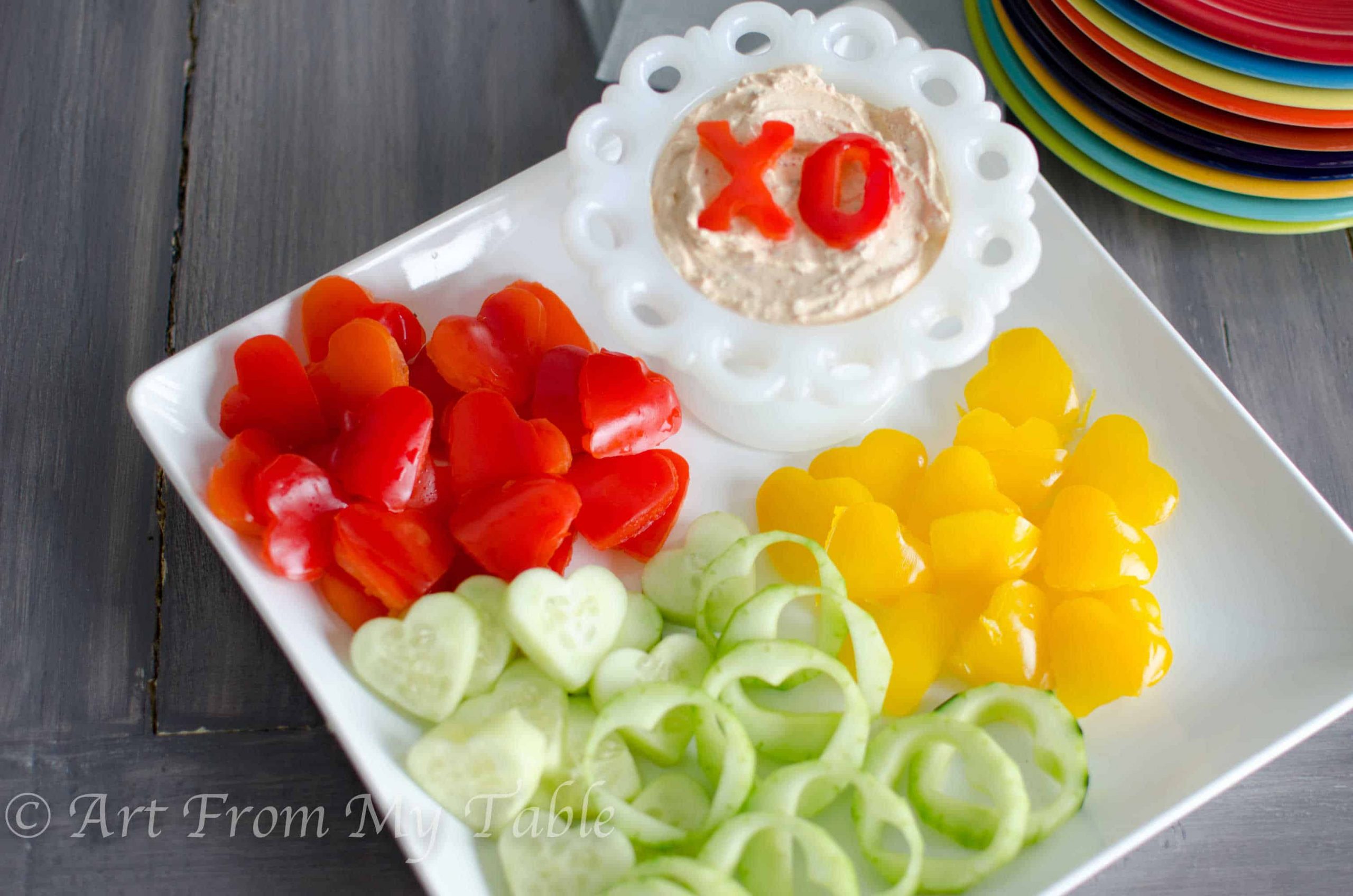 Heart shaped red and yellow bell peppers, and cucumbers on a plate with homemade veggie dip, topped with an 'x' and 'o' shaped bell pepper.