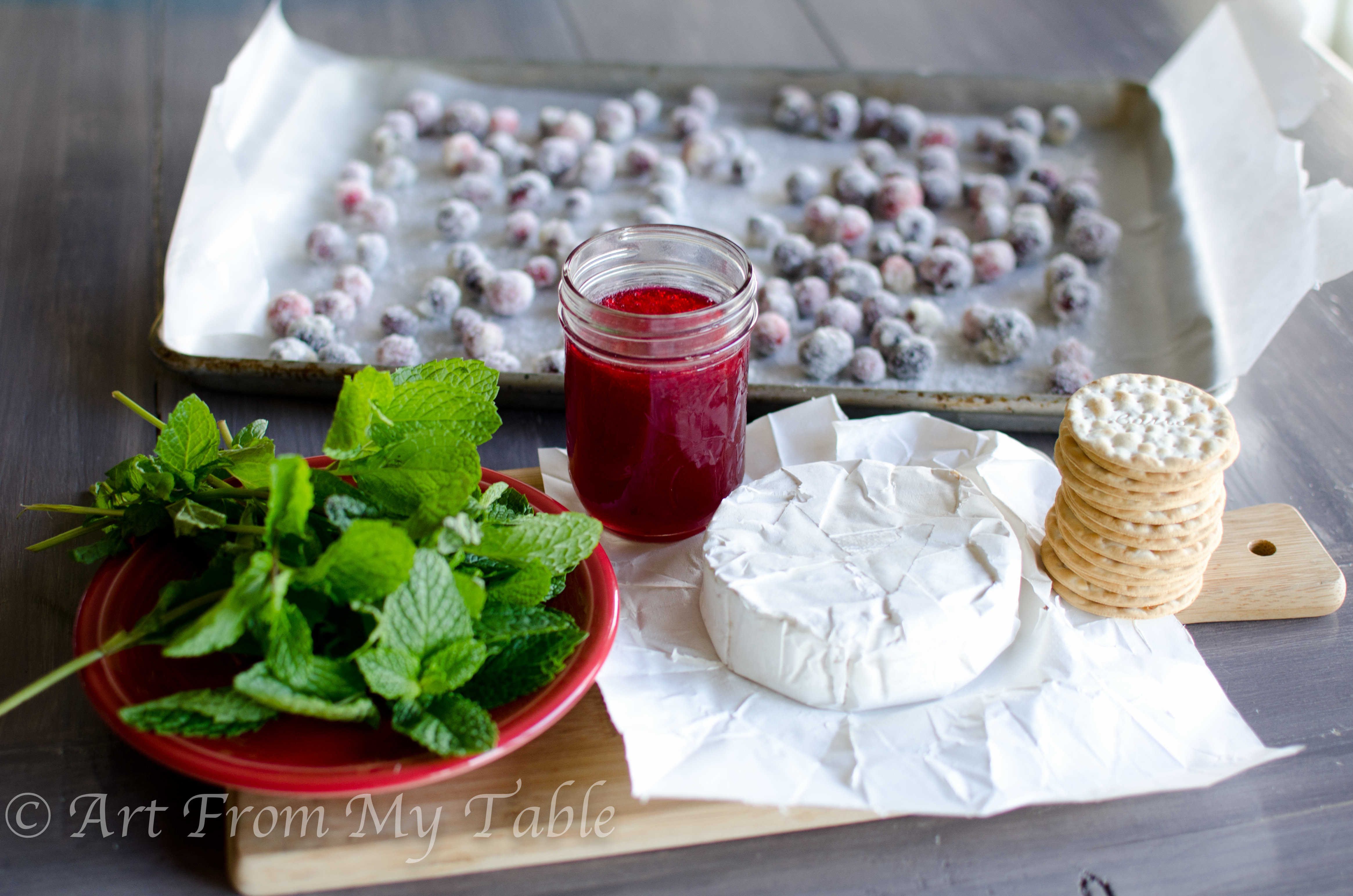Ingredients for cranberry brie bites: crackers, brie, cranberry sauce, sugared cranberries, and fresh mint leaves.