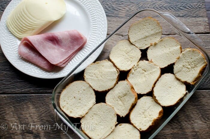 Bottom half of buns in a casserole dish next to a plate of deli ham and provolone cheese.