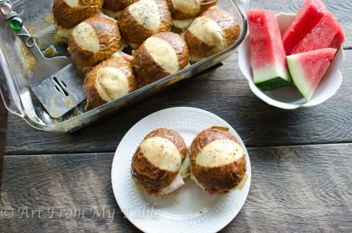 Baked ham and cheese soft pretzel sliders on a plate with watermelon and the pan of sliders in the background