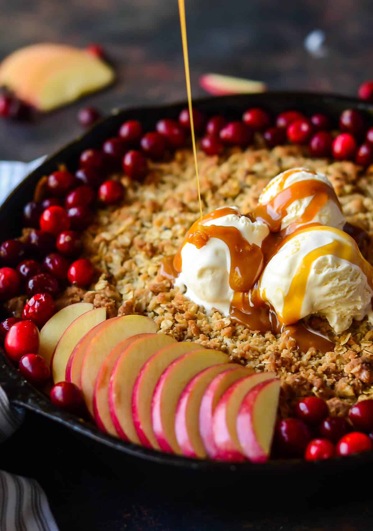 caramel sauce being drizzled over maple apple crisp topped with fresh cranberries, fresh apple slices and vanilla ice cream