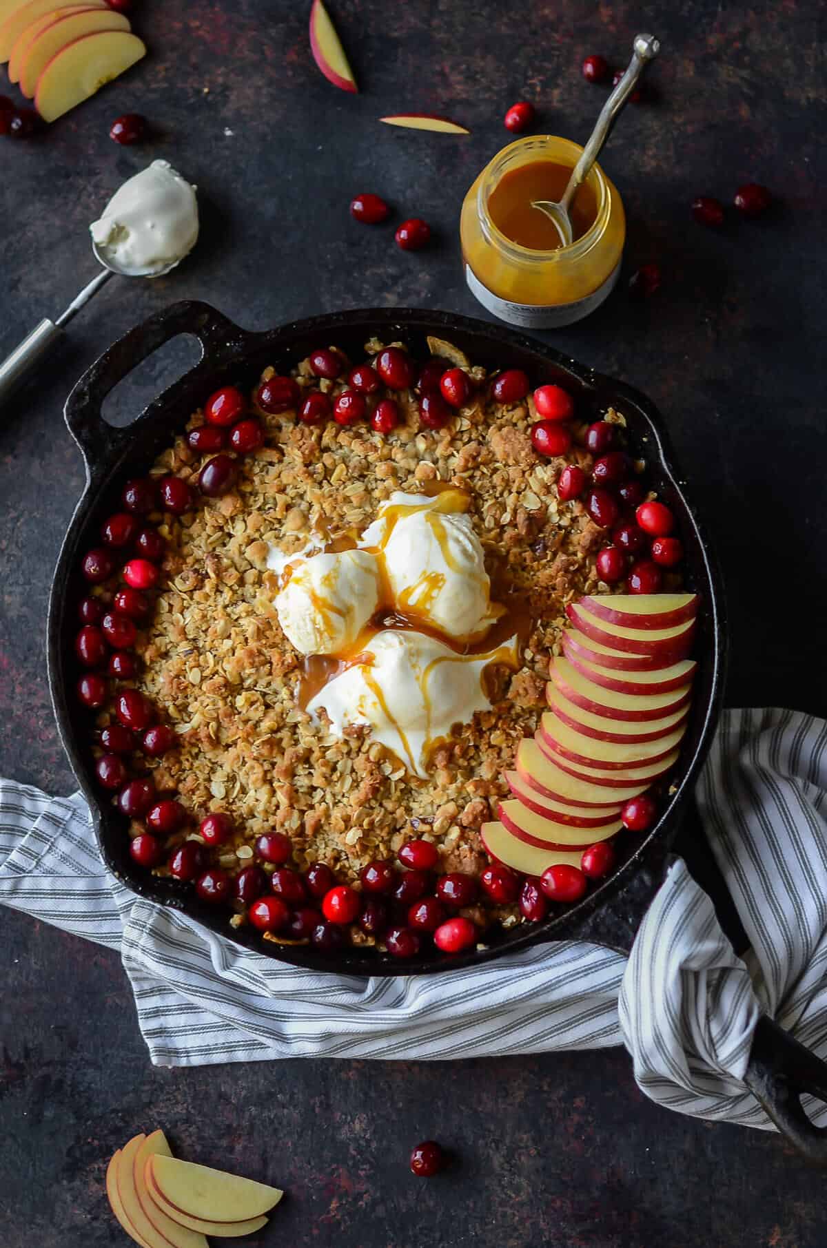 cast iron skillet filled with maple apple crisp and topped with fresh cranberries, apple slices and vanilla ice cream drizzled with caramel sauce