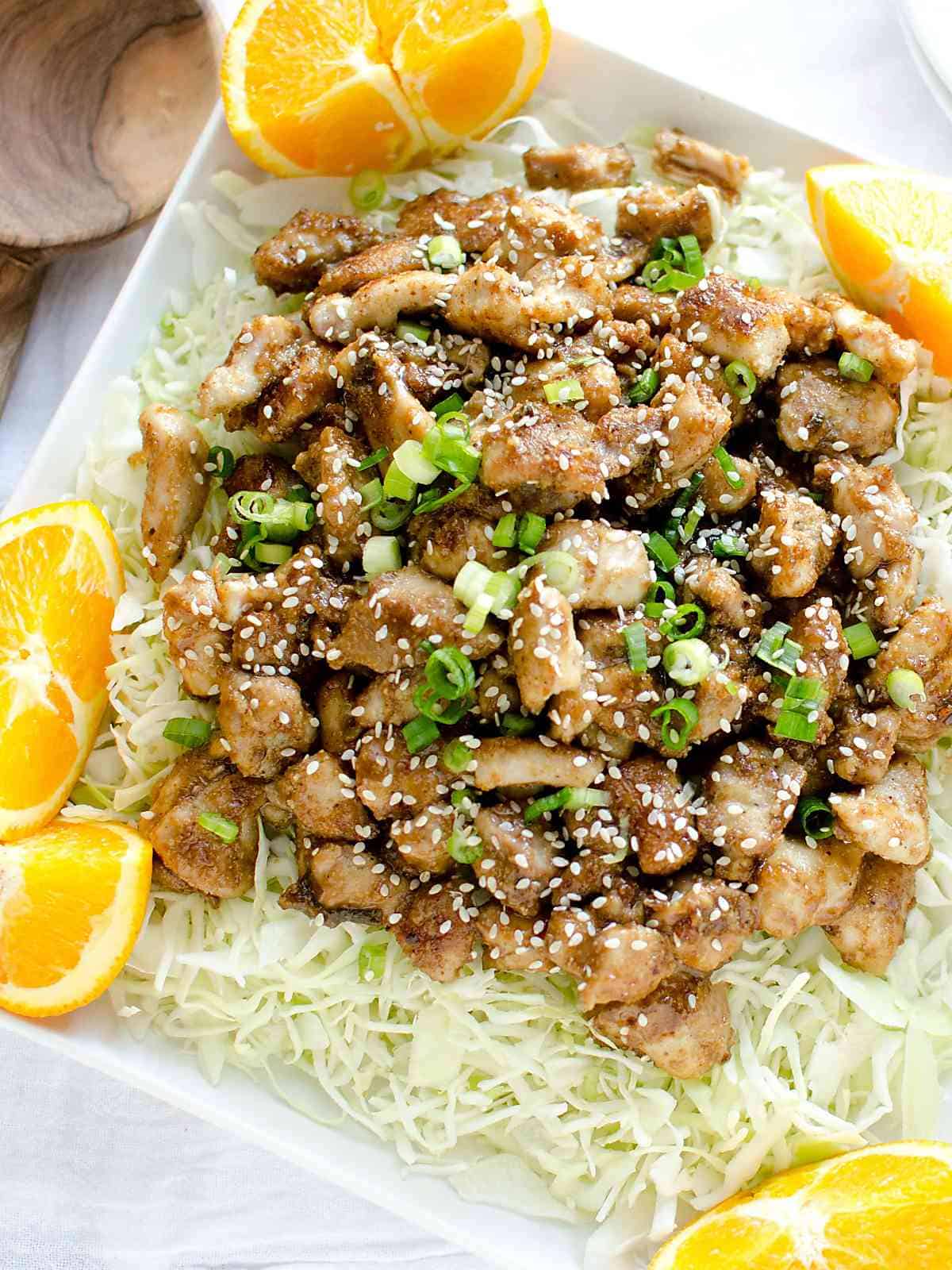 platter of homemade orange chicken served over a bed of cabbage, garnished with orange slices, green onions and sesame seeds.