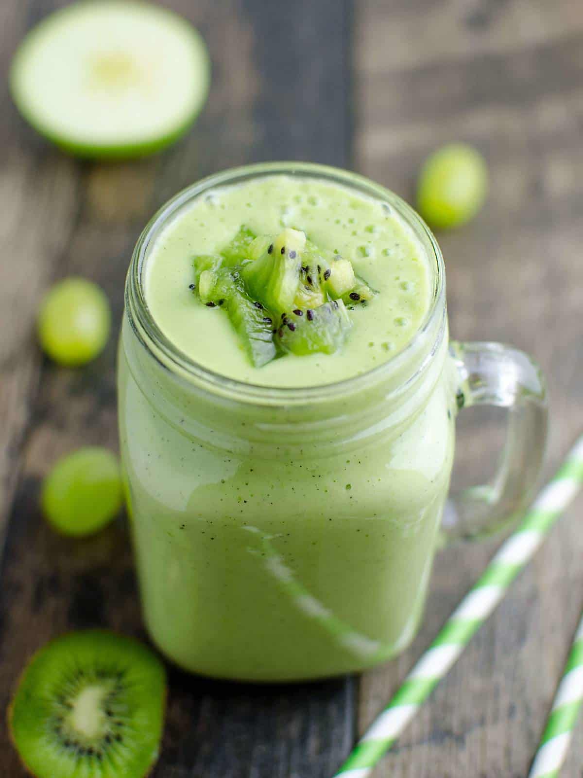 glass jar mug filled with a green smoothie made of fruit and vegetables garnished with chunks of kiwi on top.