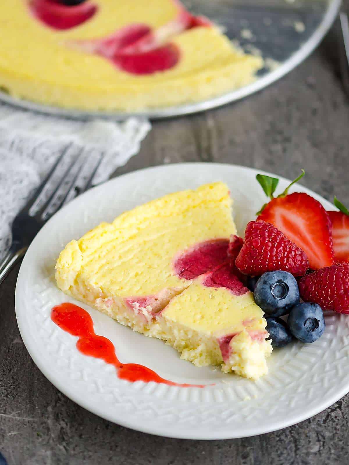 slice of 'lighter' cheesecake on a plate garnished with fresh strawberries, blueberries, and raspberries, drizzles with strawberry sauce. 