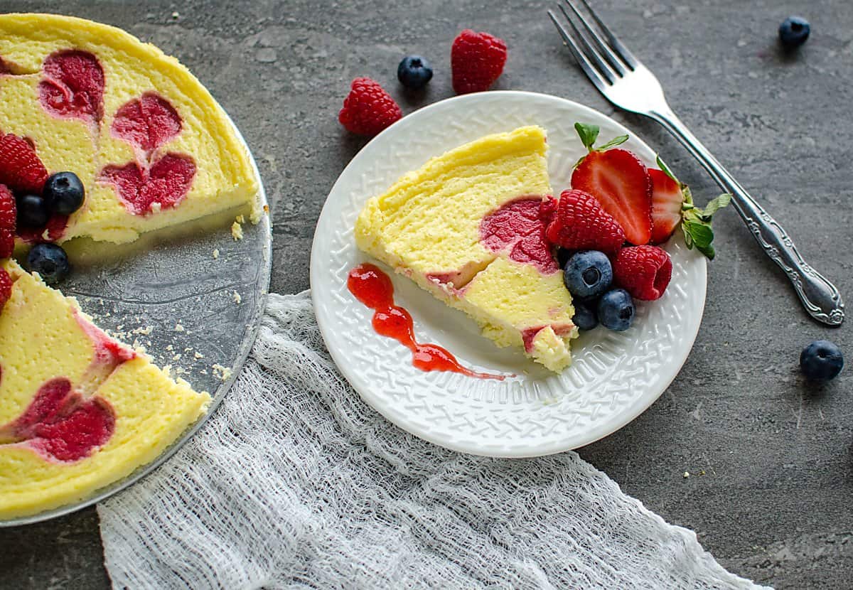 slice of cheesecake garnished with fresh berries and strawberry sauce, sitting next to the whole cheese cake minus the slice. 