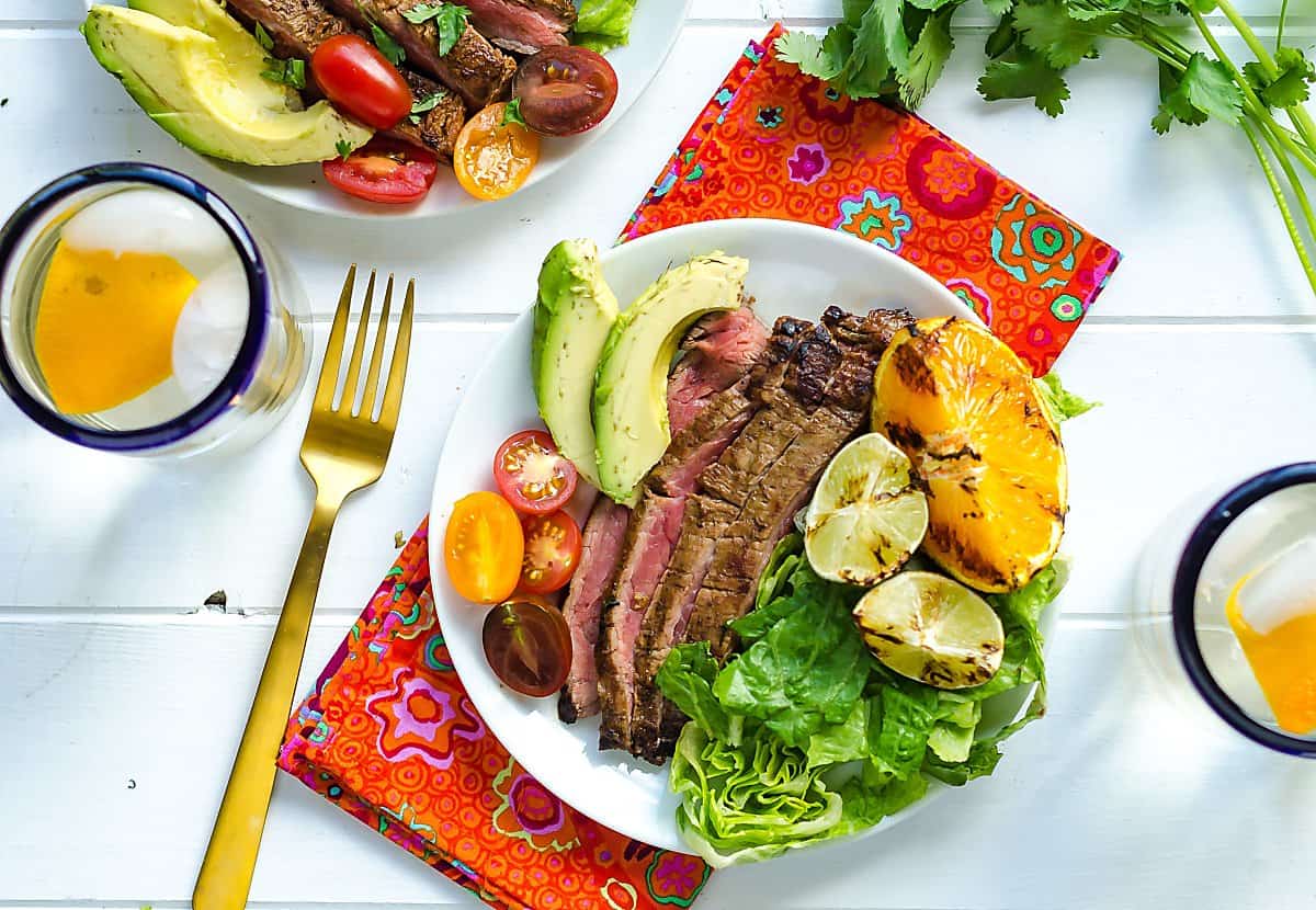 grilled Carne asada on a plate with lettuce, avocado, tomato, orange and lime wedges.