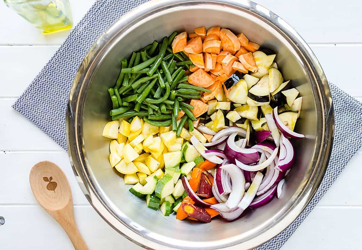 sections of cut up vegetables in a stainless steel bowl, sweet potatoes, green beans, summer squash, zucchini, carrots, red onions, eggplant