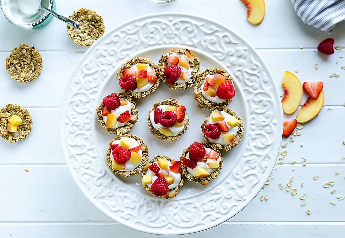 white round platter of coconut granola cups filled with yogurt, peaches, and raspberries. a few cups not filled yet, sliced peaches on the side
