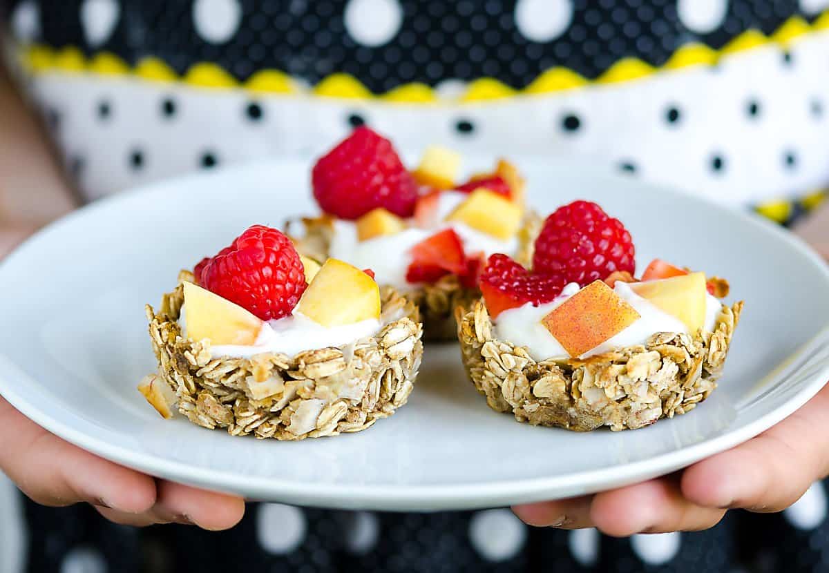 three homemade granola cups filled with yogurt, peaches and raspberries on a plate being held by someone in a polka dot apron