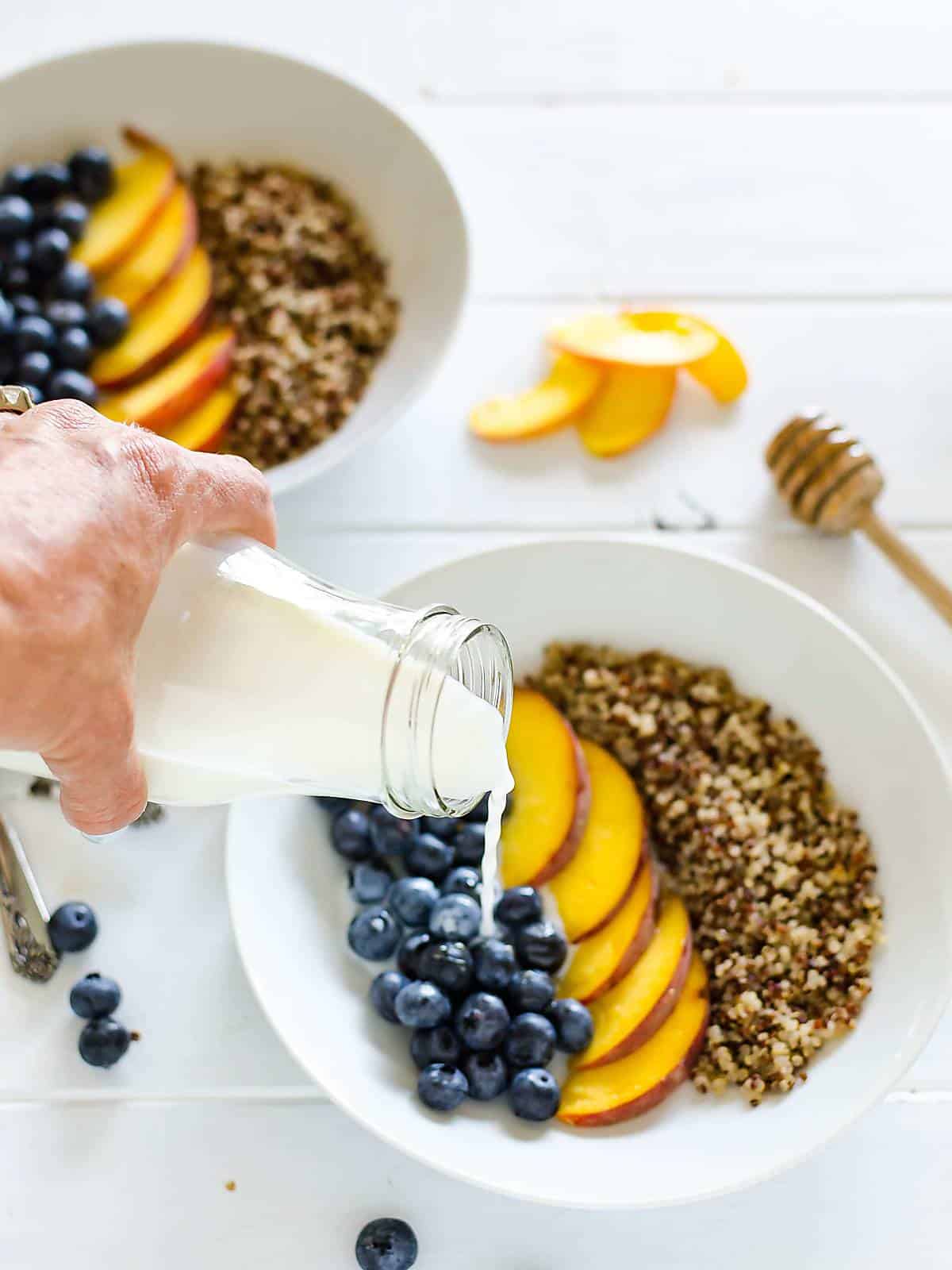 a hand pouring milk over a bowl of blueberries, peaches, and quinoa. another bowl in the back ground, a few sliced peaches on the table and a honey dipper