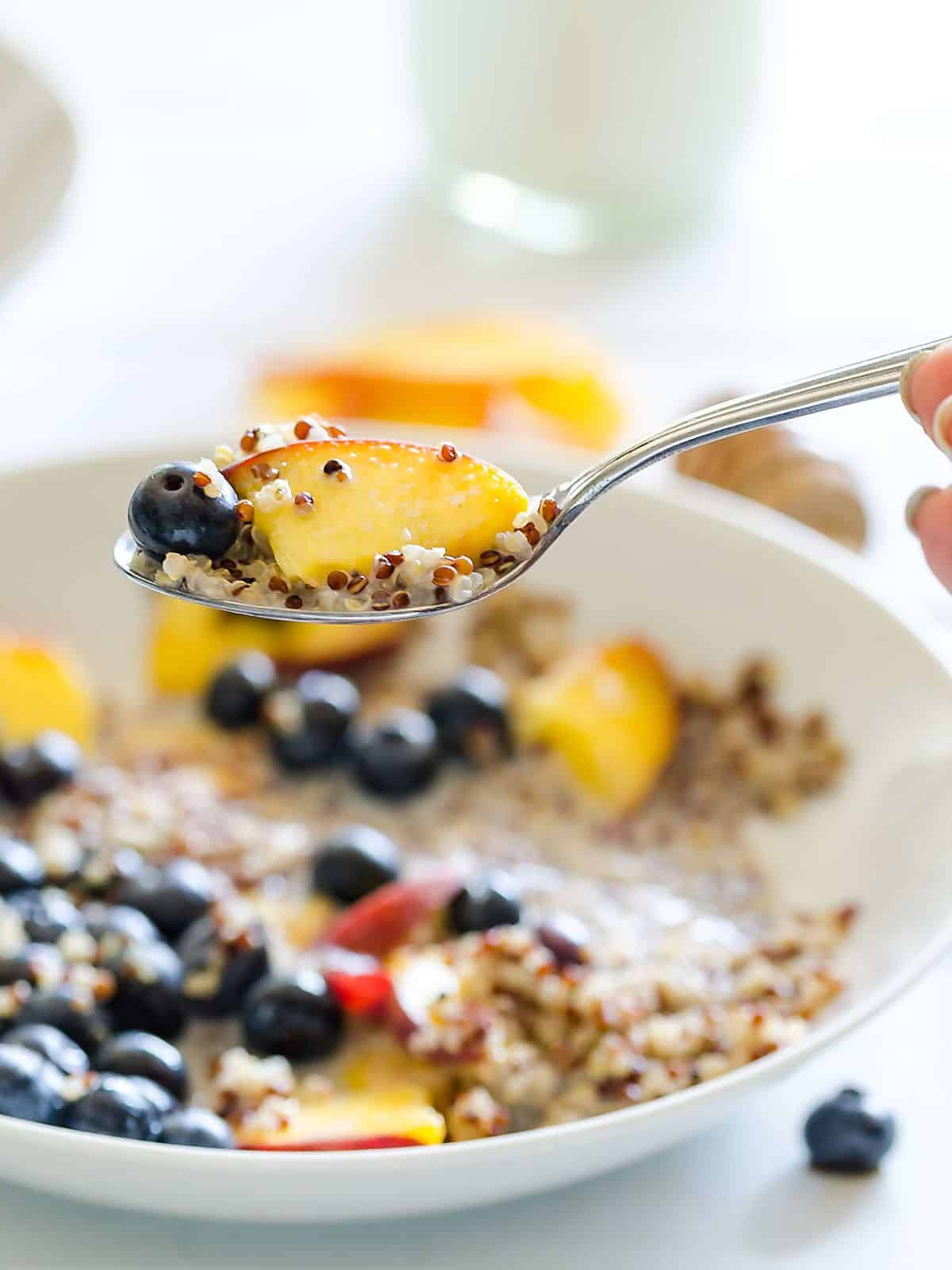 a spoon with quinoa, blueberries, a peach and milk being held above a bowl of quinoa and fruit and milk