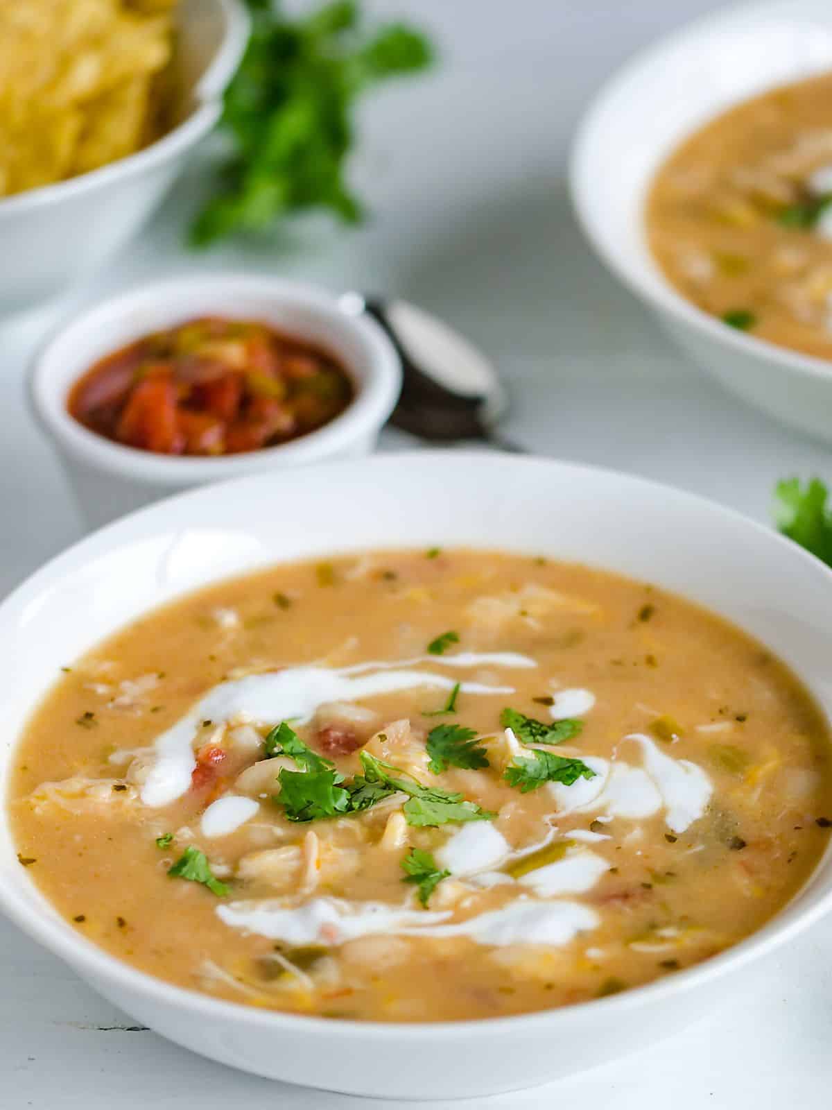 2 bowls of white chicken chili garnished with swirls of sour cream and cilantro served with chips and salsa