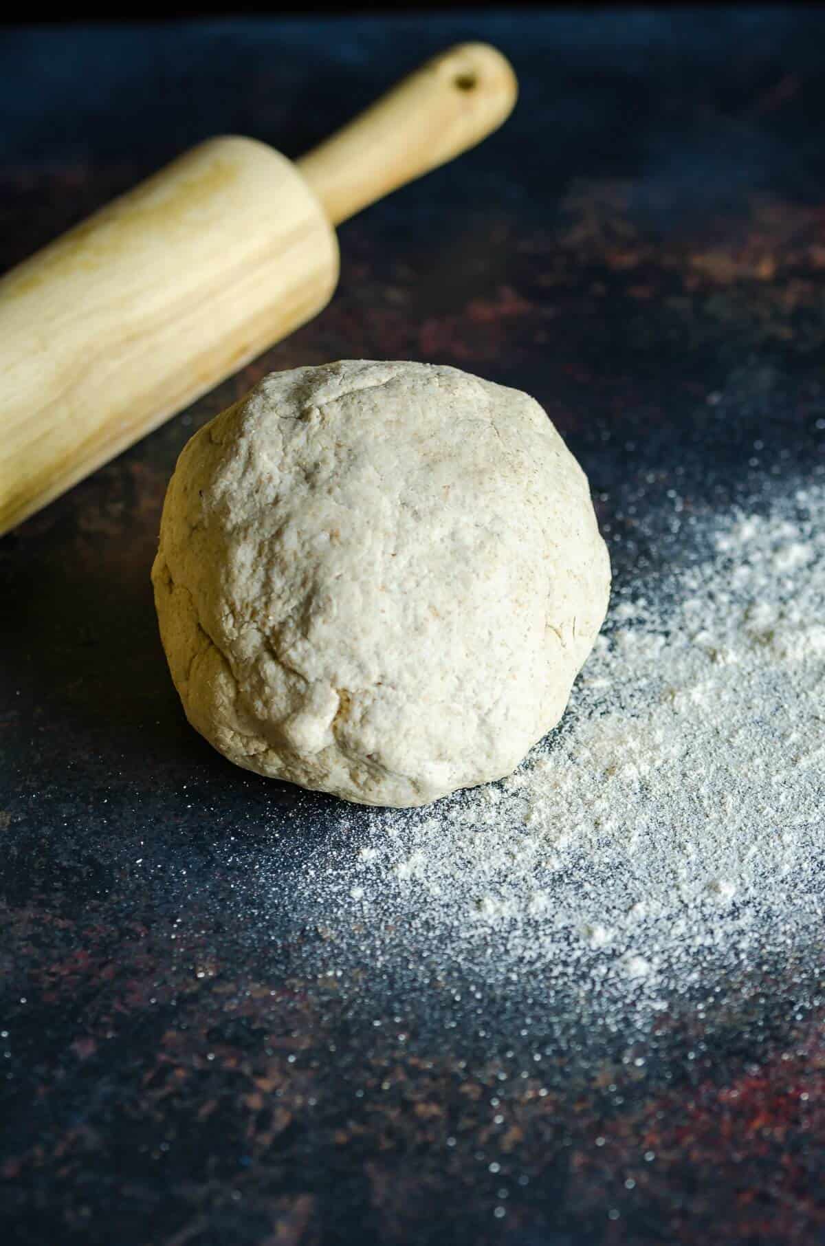 whole wheat pizza dough ball on a dark surface on flour rolling pin in the background 