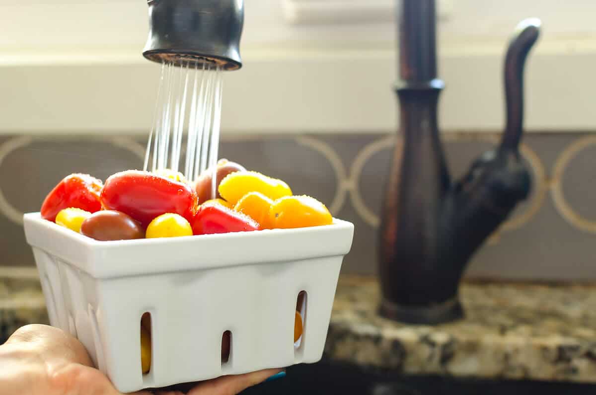 quart of red, orange, and yellow grape sized tomatoes being held under a faucet of running water.