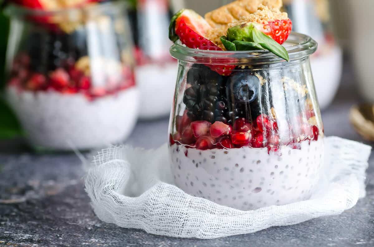 clear glass jar filled with greek yogurt chia pudding, layered with pomegranate arils, fresh berries, and belVita breakfast biscuits, a few prepared ones in the back ground.