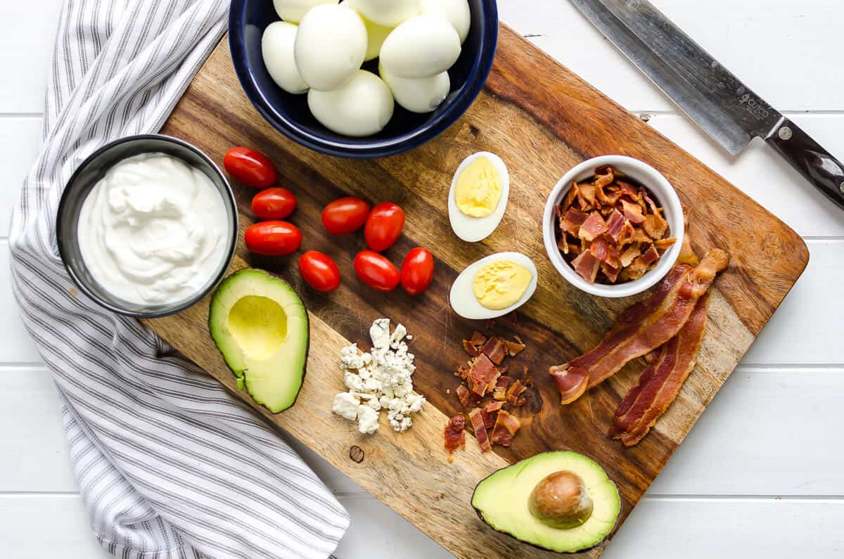 beautiful wood cutting board with cobb egg salad ingredients laid out, dark bowl full of whole hard boiled eggs, glass container of yogurt, bacon, grape tomatoes, and halved avocado
