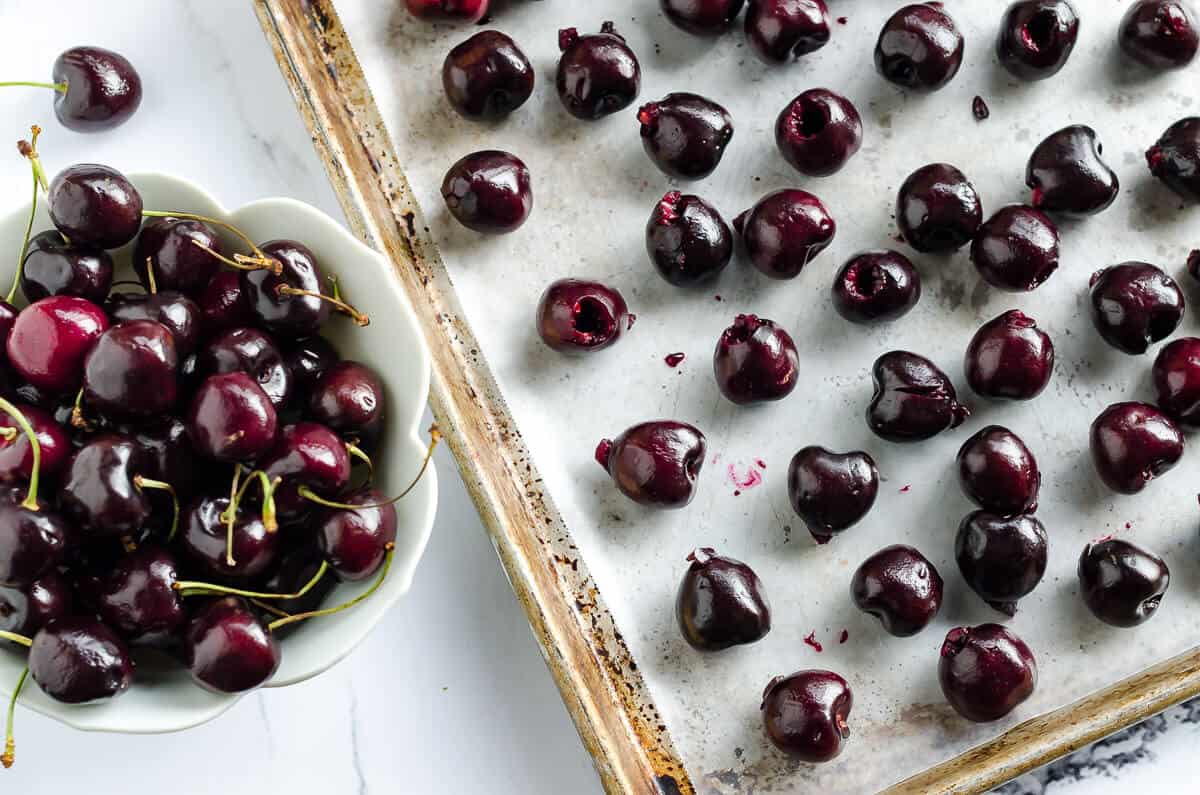 bowl of fresh sweet cherries next to a sheet pan with pitted cherries spread out for how to freeze cherries
