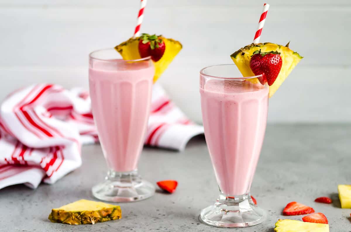 Two milkshake glasses filled with a strawberry pineapple smoothie, garnished with a slice of pineapple and a strawberry.