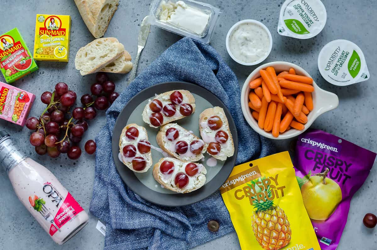 plate full of goat cheese crostini surrounded by healthy lunch options: carrots, ranch, sunmaid raisins, cluster of grapes and crispy fruit packages