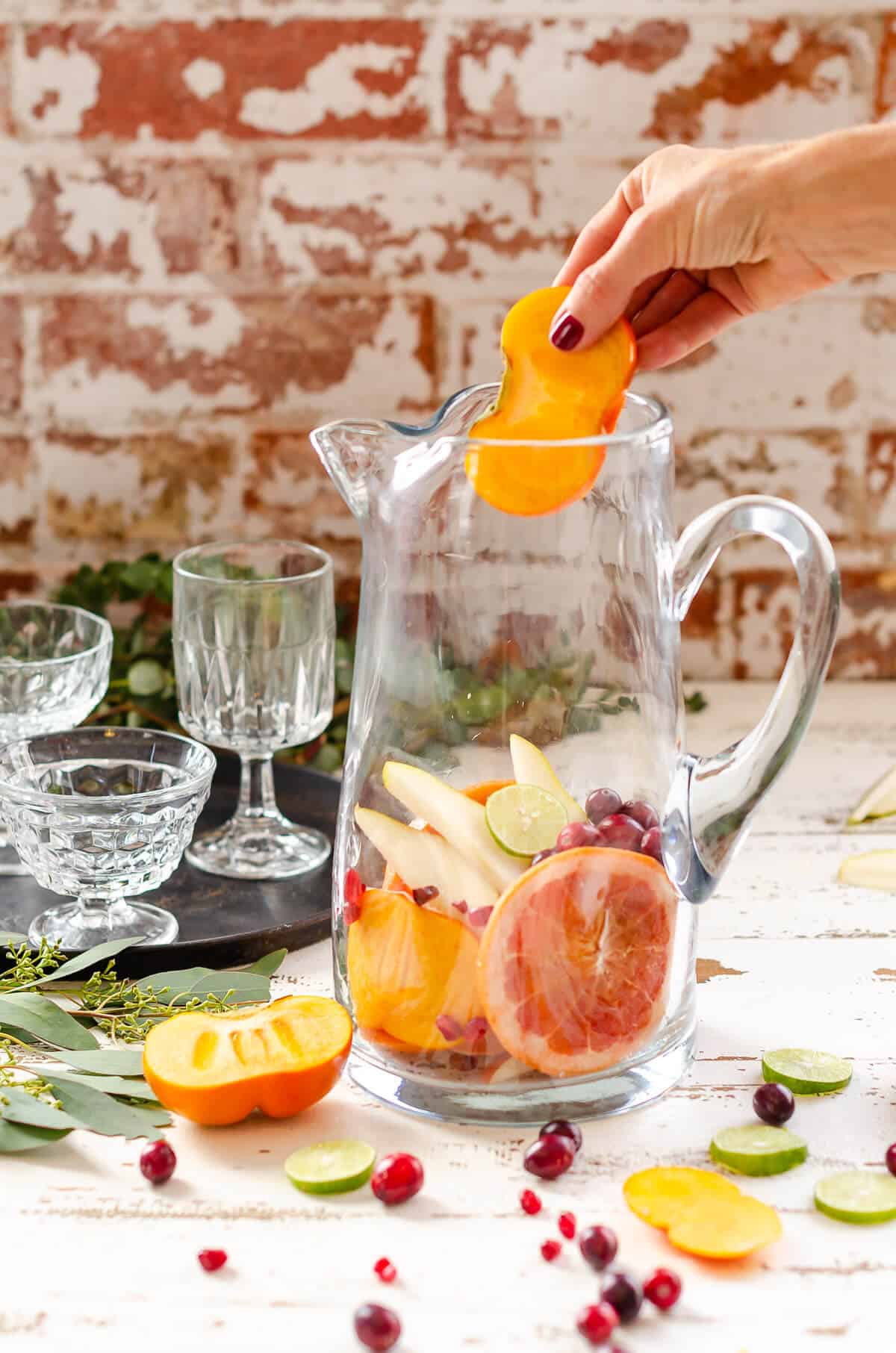 hand adding a slice of persimmon to a clear glass pitcher filled with fresh grapefruit, pears and cranberries for a Sangria Mocktail
