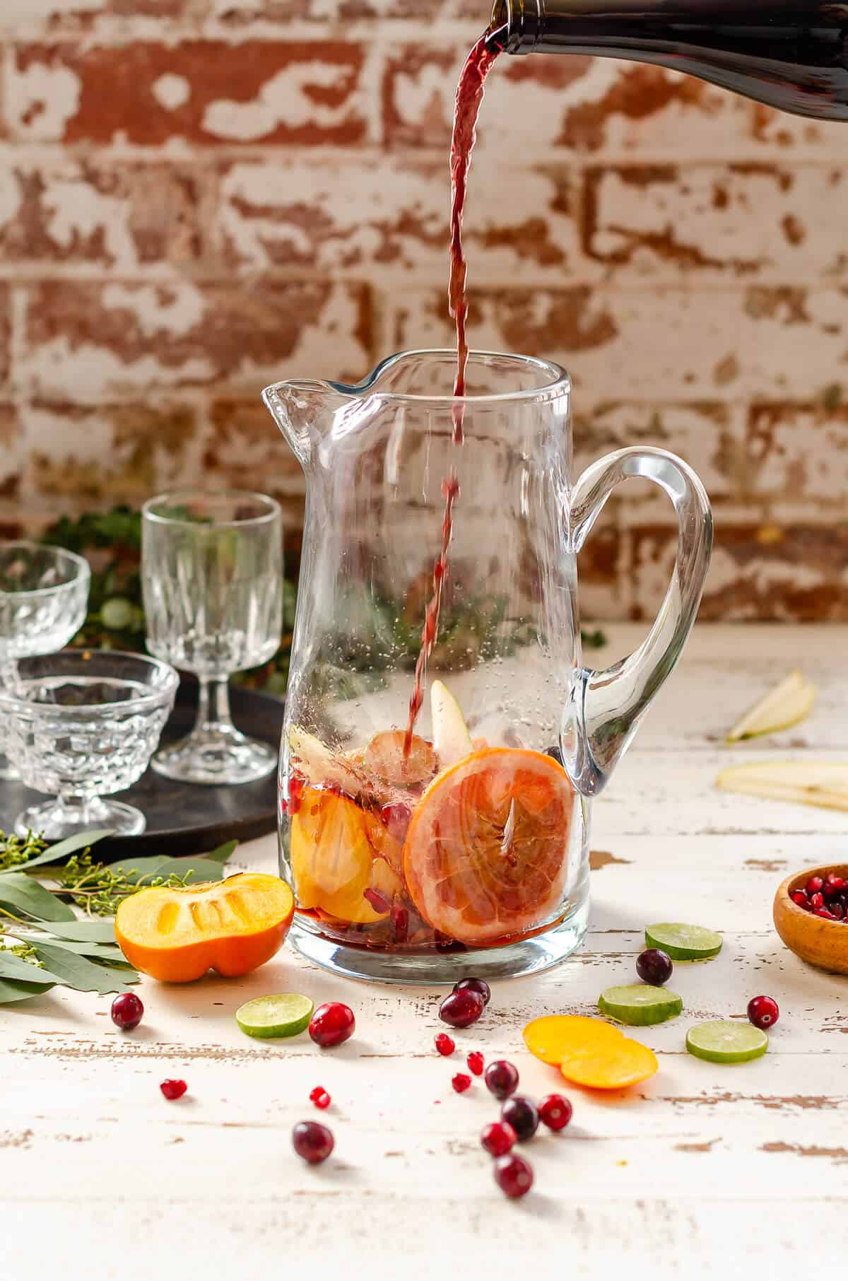 clear glass pitcher with grapefruit, persimmon, pears, and cranberries in the bottom, with red non-alcoholic wine being poured in.