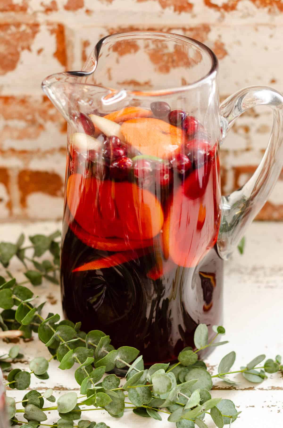 Clear glass pitcher filled with non alcoholic red sangria mocktail, fresh grapefruit, oranges, persimmon, and cranberries. greens surrounding the pitcher