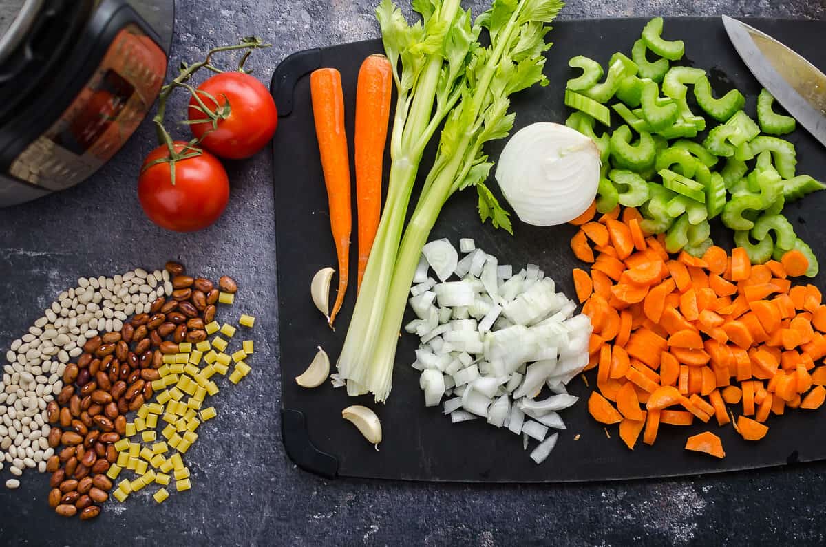 cutting board with chopped carrots, celery and onions along side dried beans, pasta and tomatoes