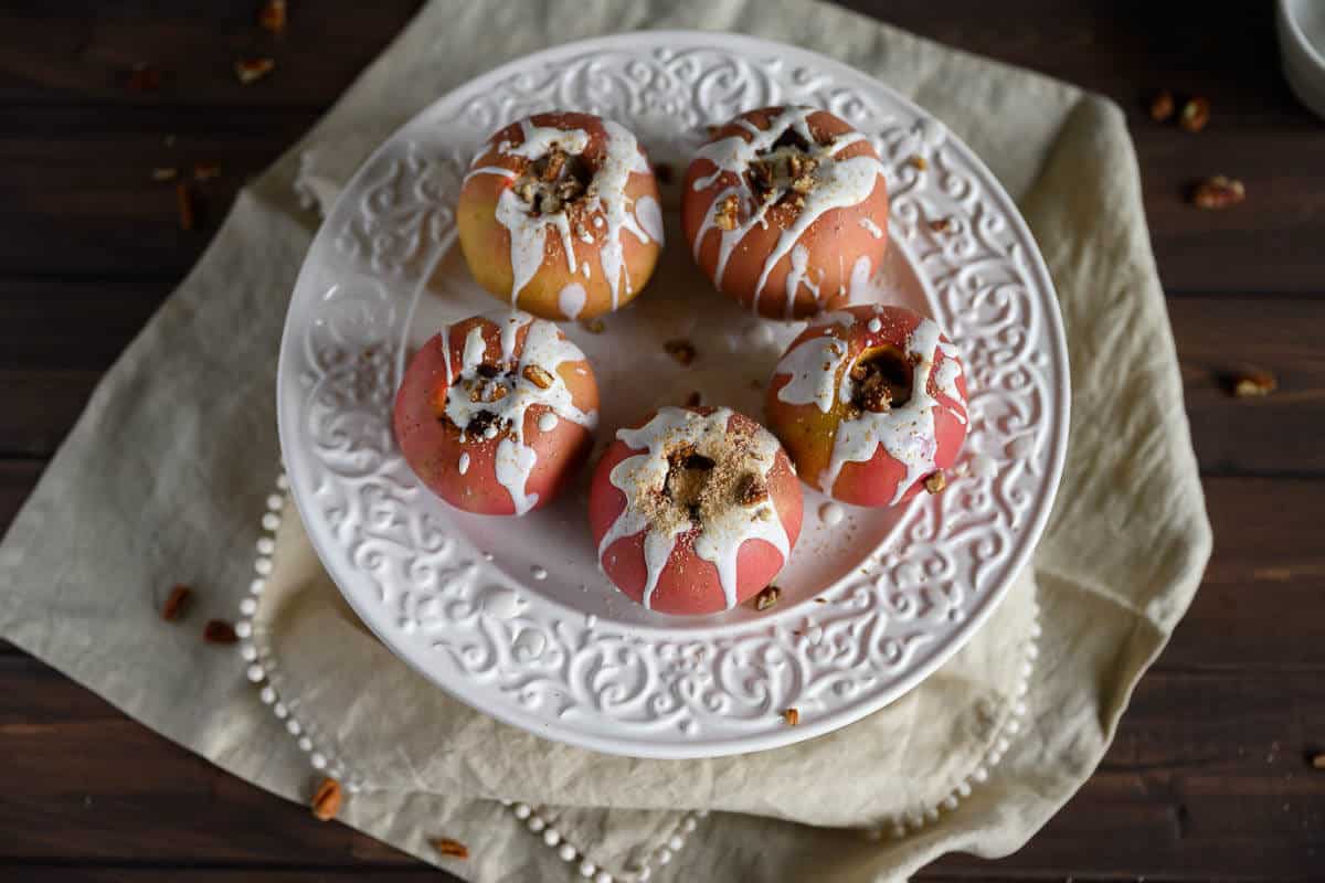 5 healthy baked apples on a white platter drizzled with yogurt sauce, chopped pecans and nutmeg