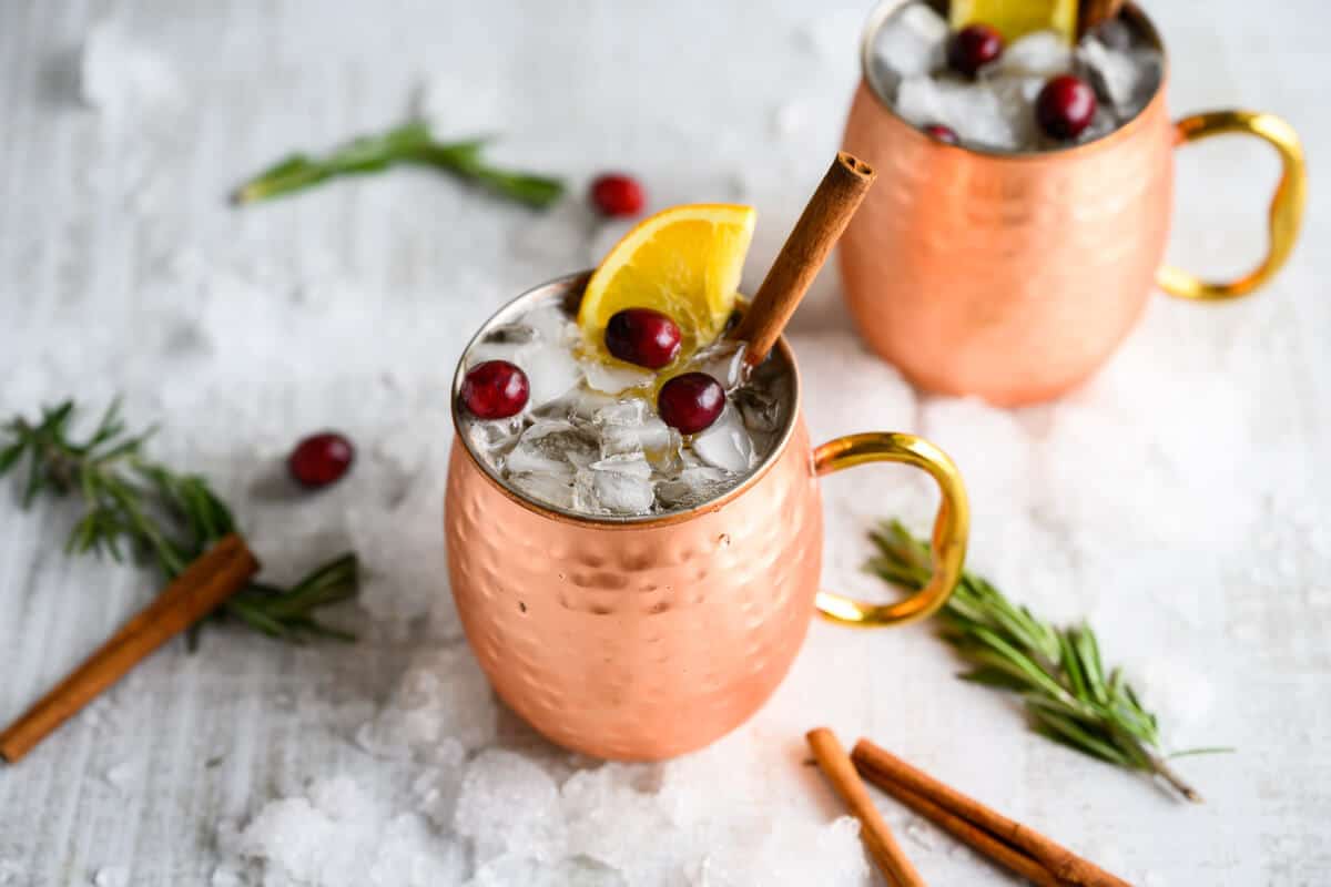 two copper mugs filled with cranberry moscow mule mocktail, surrounded by snow, cinnamon sticks and rosemary