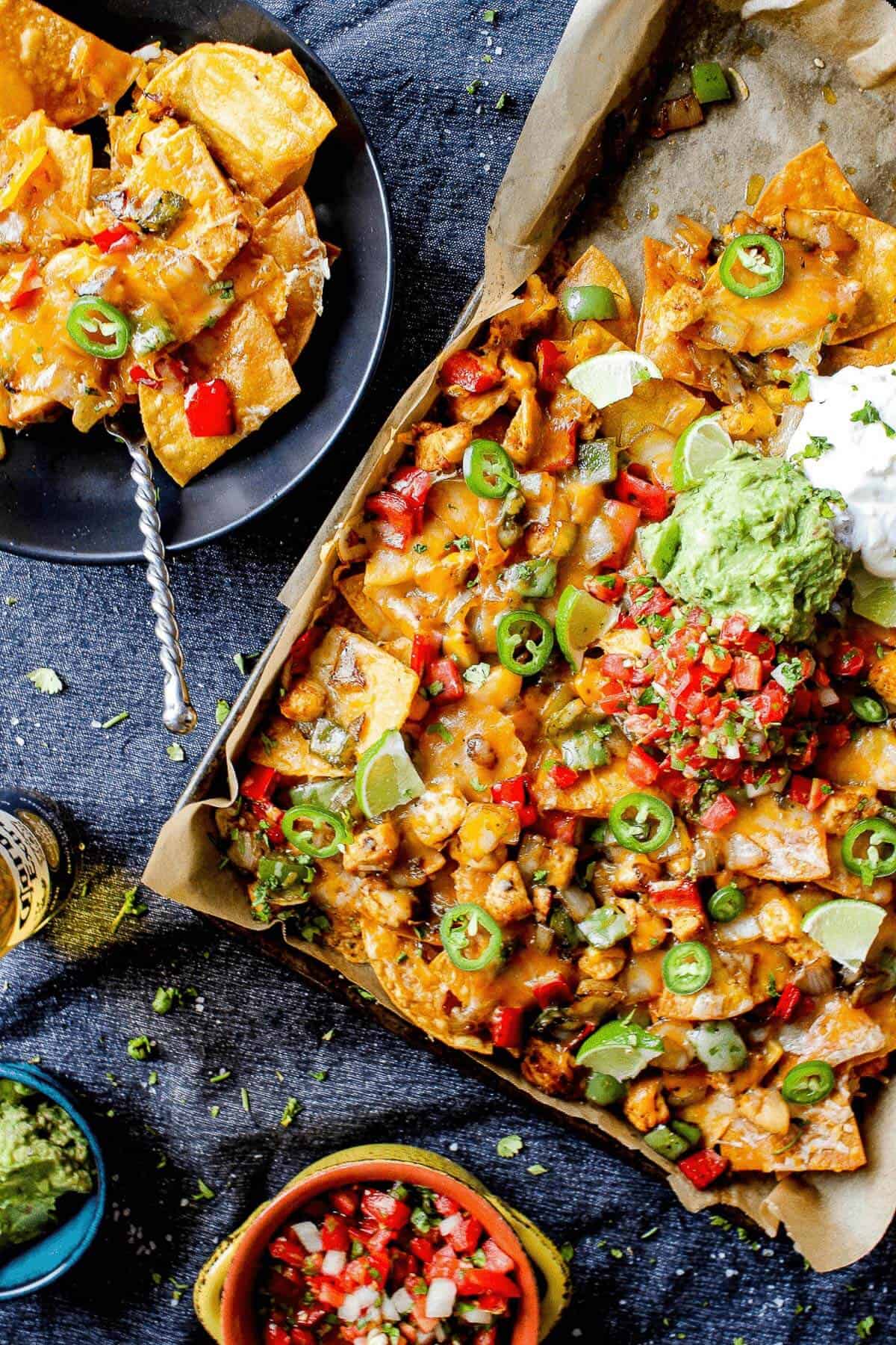 sheet pan full of chicken fajita nachos alongside garnishes of salsa, guacamole, and a plate full fo nachos
