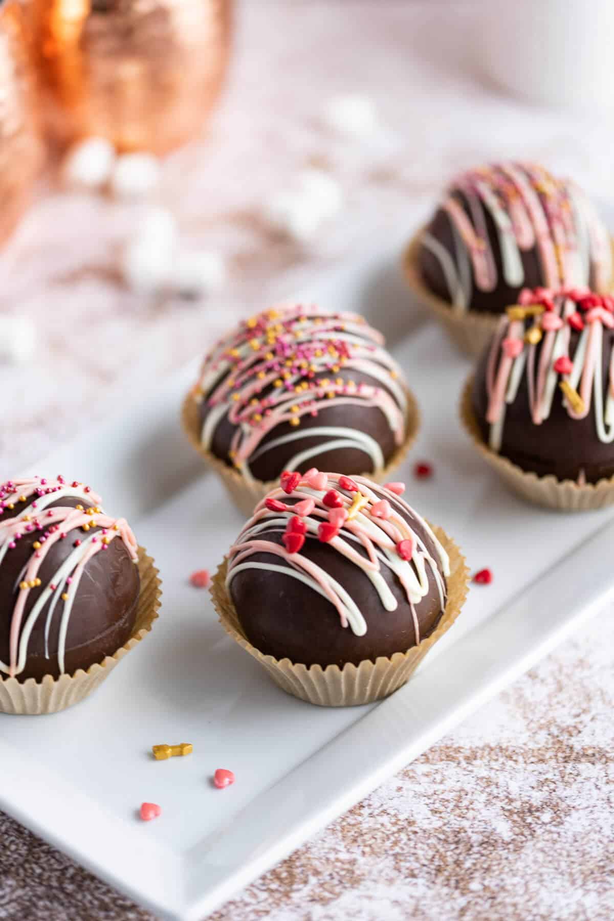 a platter of hot chocolate bombs decorated with pink and white chocolate drizzle and valentine sprinkles
