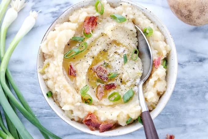 speckled bowl filled with colcannon, garnished with bacon and green onions, spoon digging in.