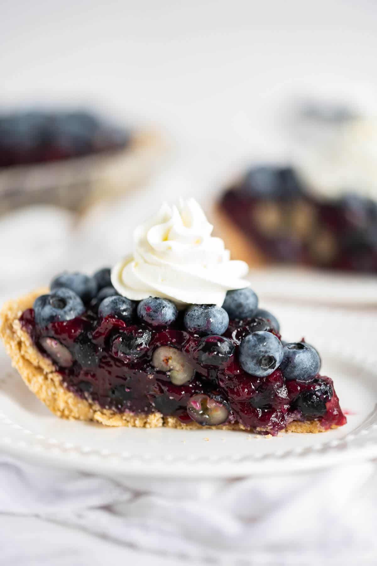 blueberry pie with graham cracker crust on a white plate topped with whipped cream