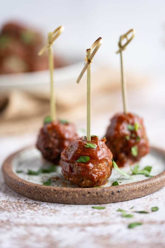 Three BBQ party meatballs on a plate with toothpicks in them and garnished with chopped parsley.