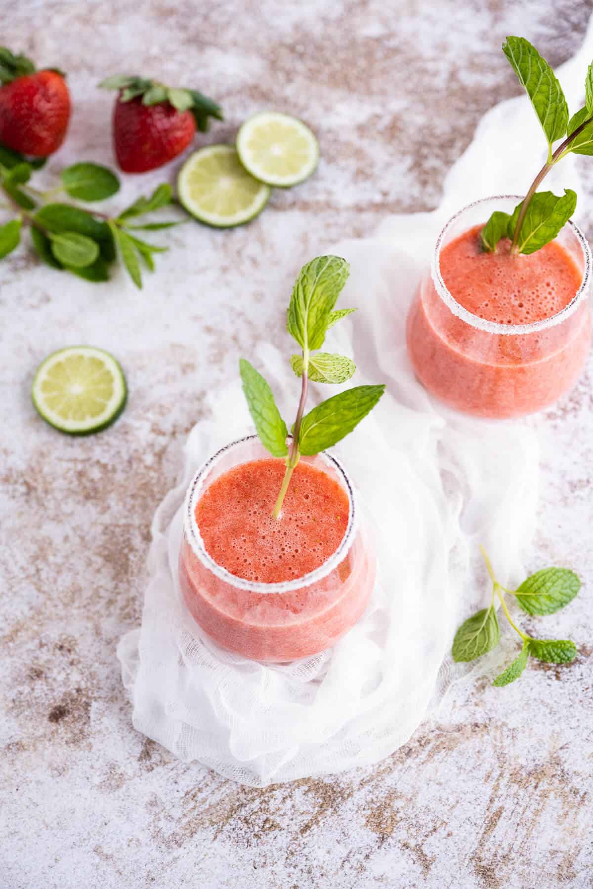 Two Strawberry Mocktails in clear stemless wine glasses garnished with fresh mint. Glass is rimmed in sugar. Sliced limes and fresh strawberries in the background.