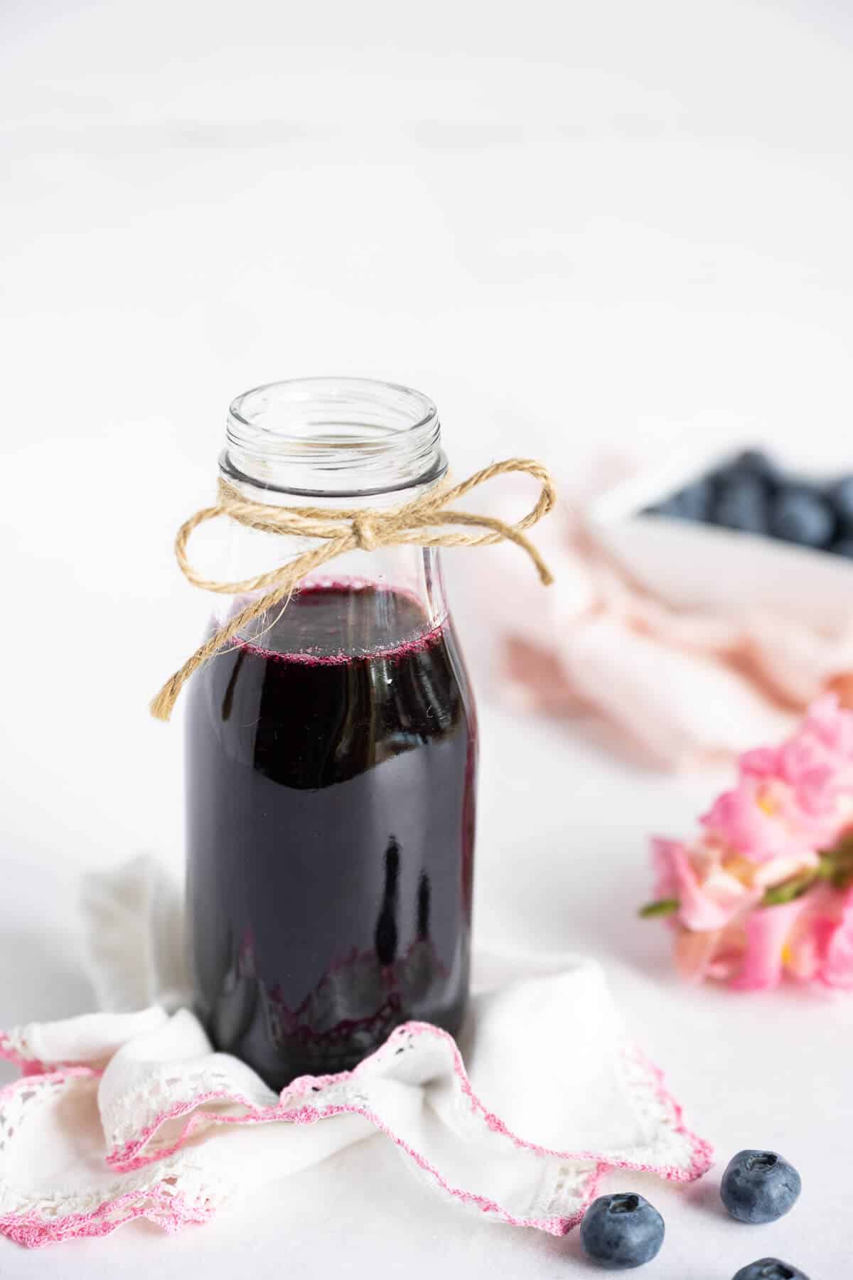 Blueberry simple syrup in a glass jar. Bowl of blueberries in the background, fresh flowers along side.