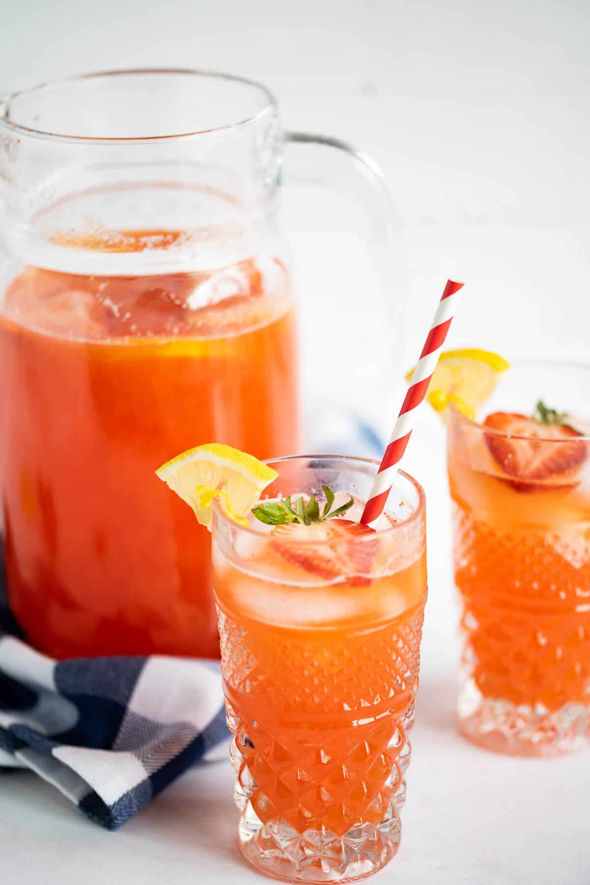 A glass of strawberry lemonade garnished with a fresh strawberry and lemon wedge. Behind the glass is another glass and a pitcher of the lemonade.