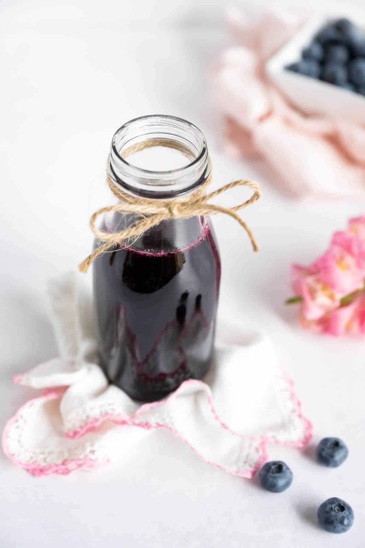 Homemade blueberry simple syrup in a milk-bottle type glass jar. A few blueberries off to the side and a bowl of fresh blueberries in the background.