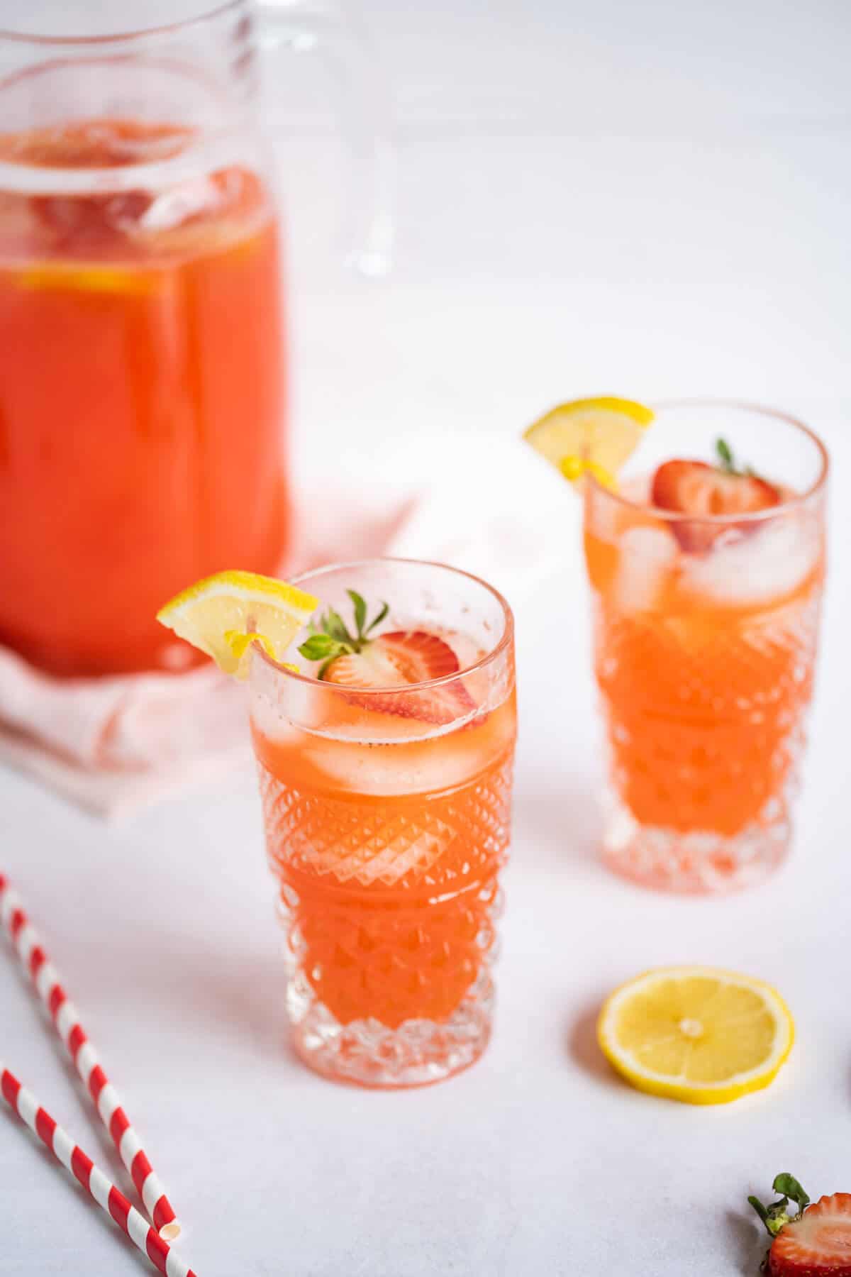 Two glasses of Strawberry Lemonade with a pitcher of lemonade behind them. The drinks are garnished with fresh strawberries and a lemon wedge. 2 straws are beside the glass.
