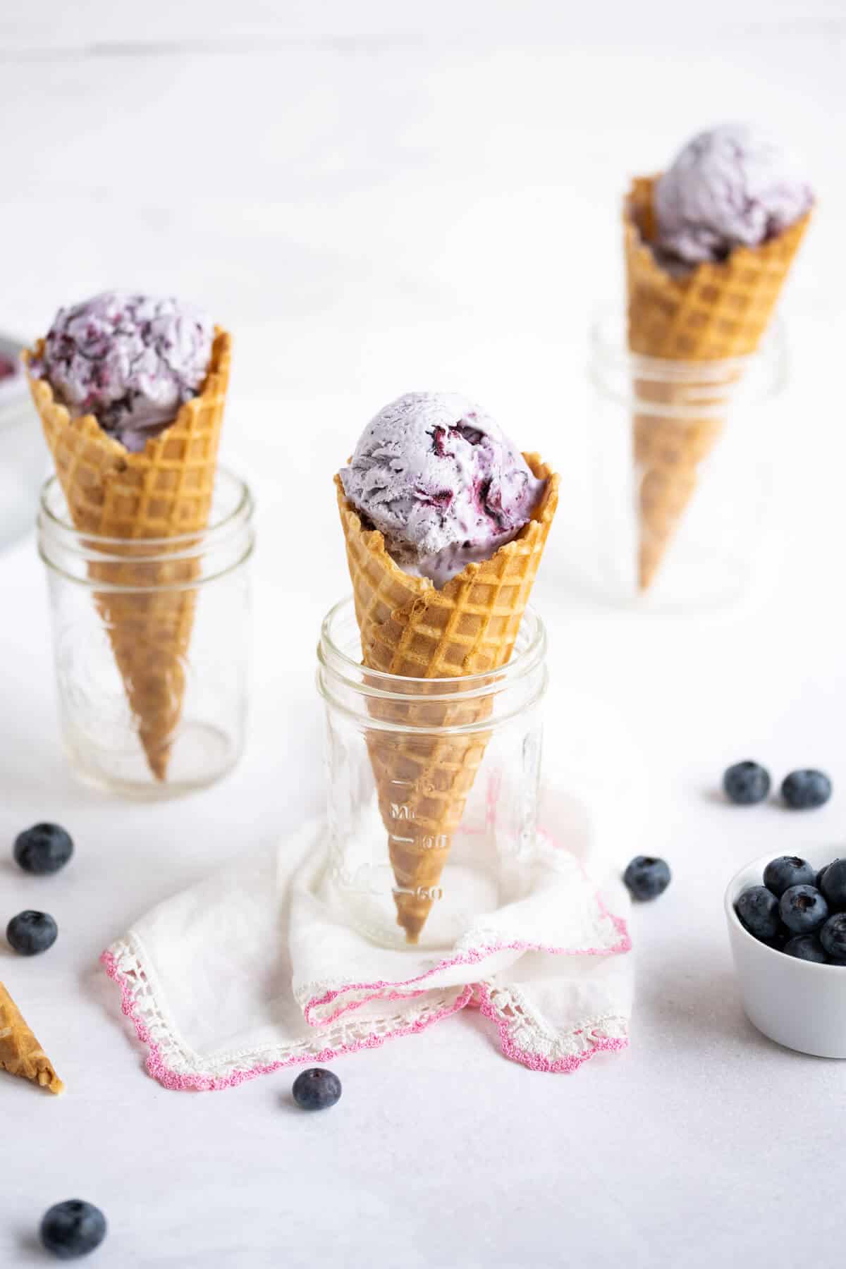 3 blueberry ice cream cones, sitting in 3 mason jars. Fresh blueberries are scattered in the foreground.