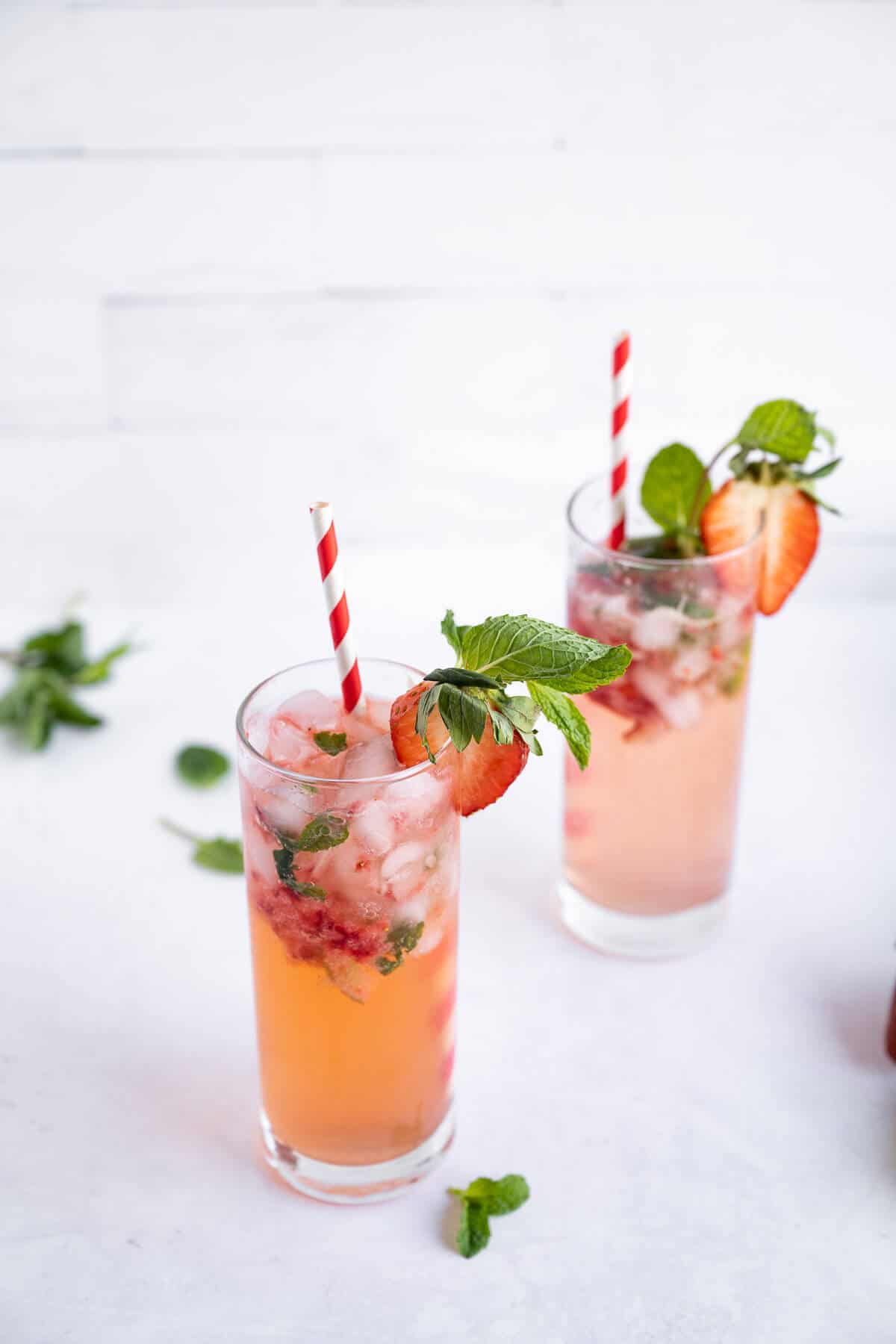 2 glasses of freshly made strawberry mojito mocktails. Drinks are garnished with fresh mint leaves and sliced strawberries. Each glass has a red and white striped straw in it.