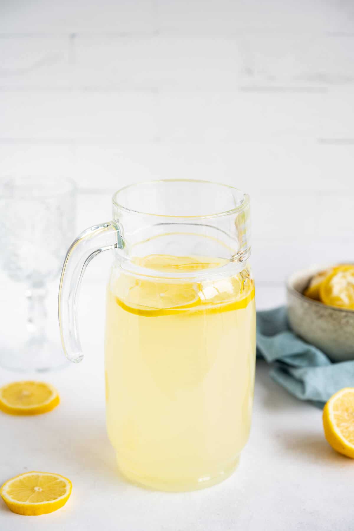 Clear glass pitcher filled with Keto Lemonade. Lemon slices laying next to the pitcher and inside.