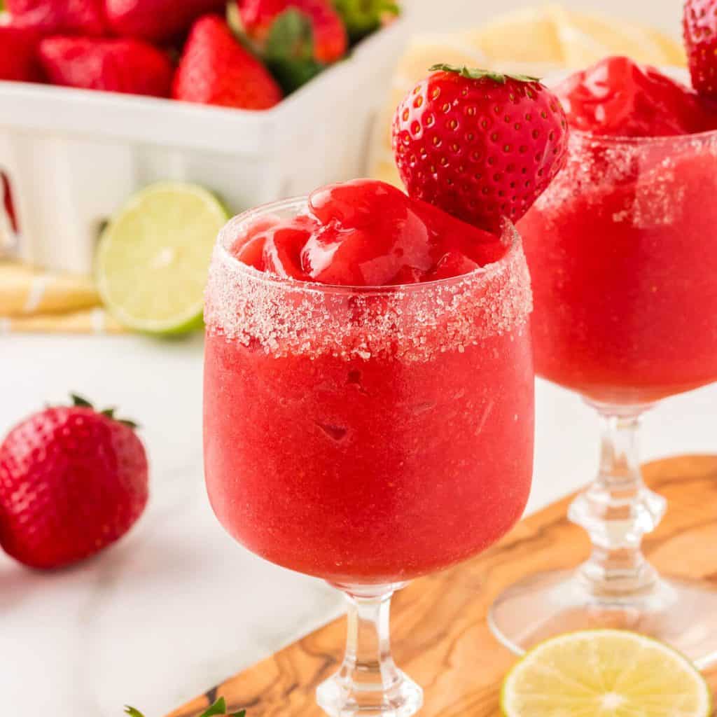 Virgin strawberry daiquiris in a sugar rimmed glass, garnished with a strawberry. A quart of strawberries is in the background, and there are a few strawberries and lime wheels scattered.