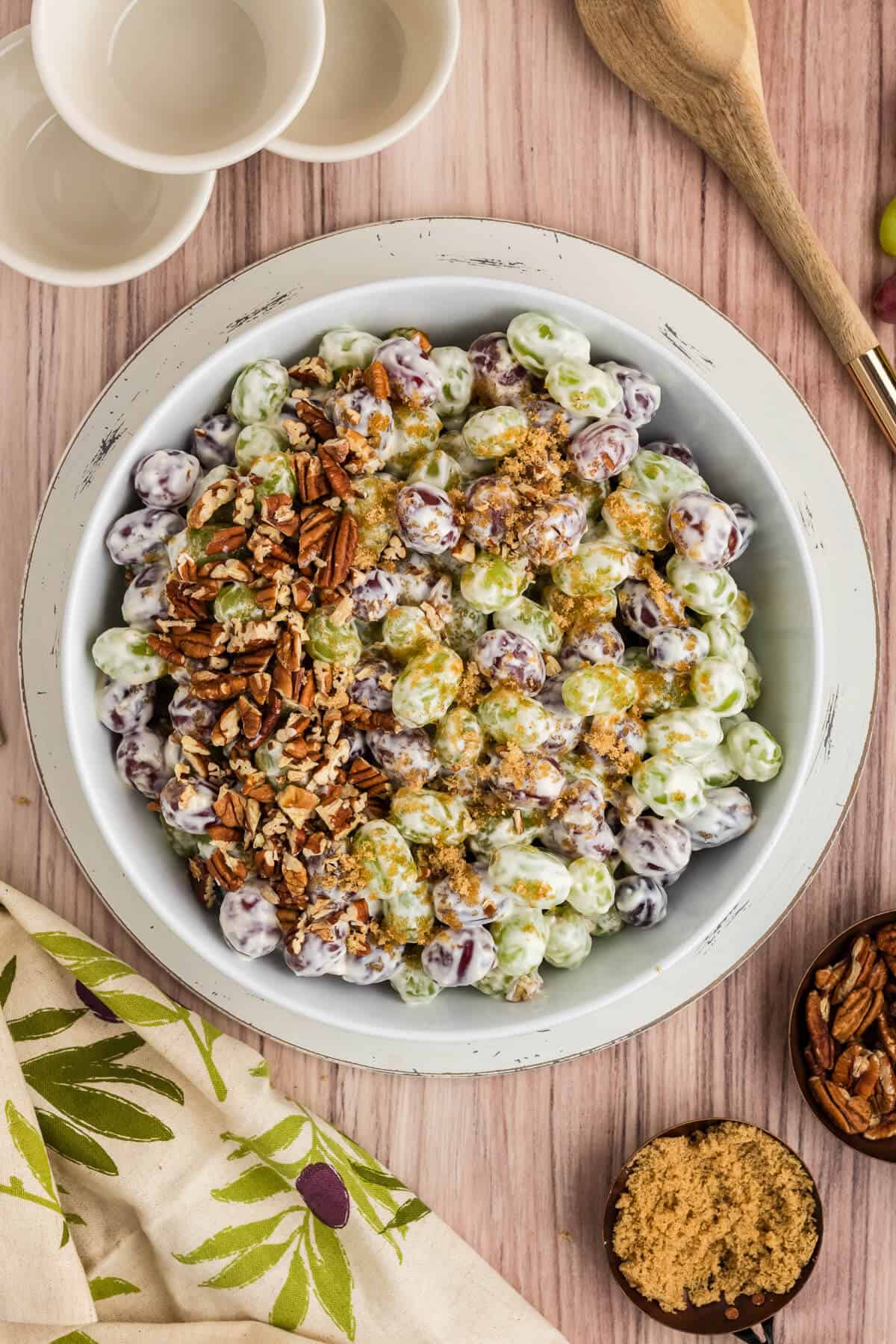 Large white bowl of Grape Salad, garnished with brown sugar and chopped pecans. Serving bowls and spoon are off to the side. A scoop of brown sugar and a scoop of pecans are also near the serving bowl.