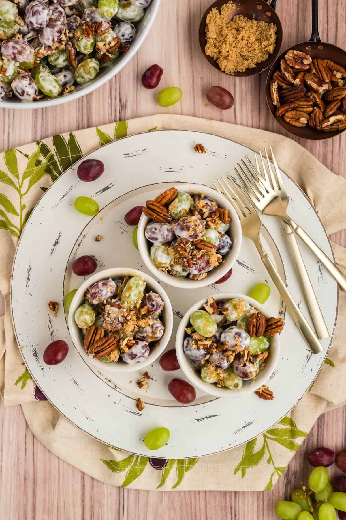 3 bowls of Chicken Salad Chick Grape Salad on. a round white platter with a few grapes scattered about. Large bowl of grape salad, scoop of brown sugar and a scoop of pecans are off to the side.