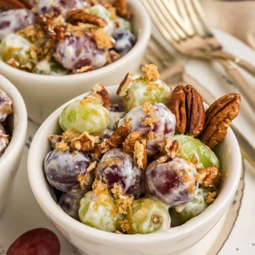 Close up picture of grape salad in a small serving bowl, garnished with brown sugar and chopped pecans. A couple of whole pecans in the corner of the bowl. There are more bowls of grape salad in the background and some gold forks.