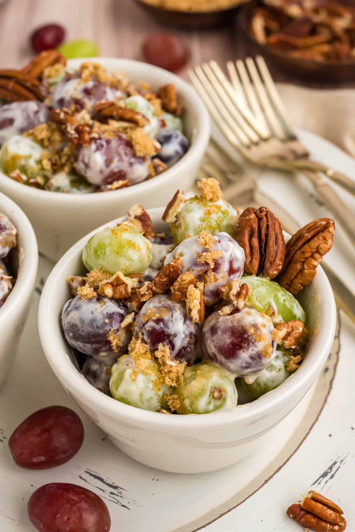 Close up picture of grape salad in a small serving bowl, garnished with brown sugar and chopped pecans. A couple of whole pecans in the corner of the bowl. There are more bowls of grape salad in the background and some gold forks.