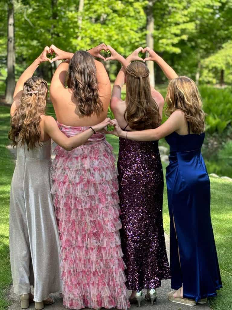 Four friends in prom dresses, from behind, making hearts with their hands.